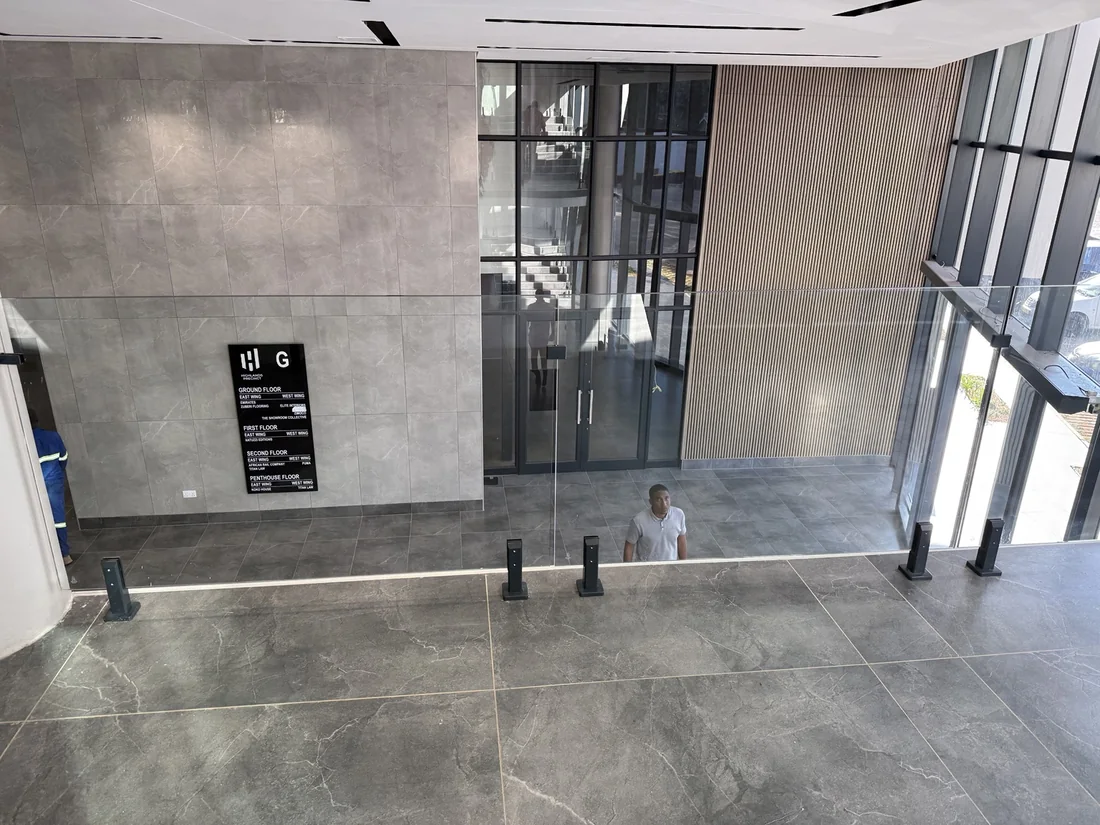 Glass balustrade with dark metal fixings overlooking a tiled atrium and tall windows, in Kingson Engineering’s supplied project photograph.