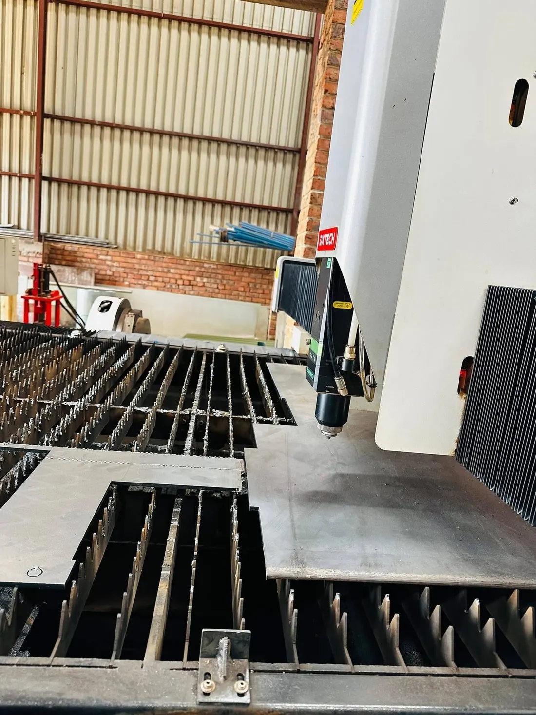 The cutting head of a fibre laser positioned over steel plate on a slatted bed, with the machine casing and workshop wall behind it.