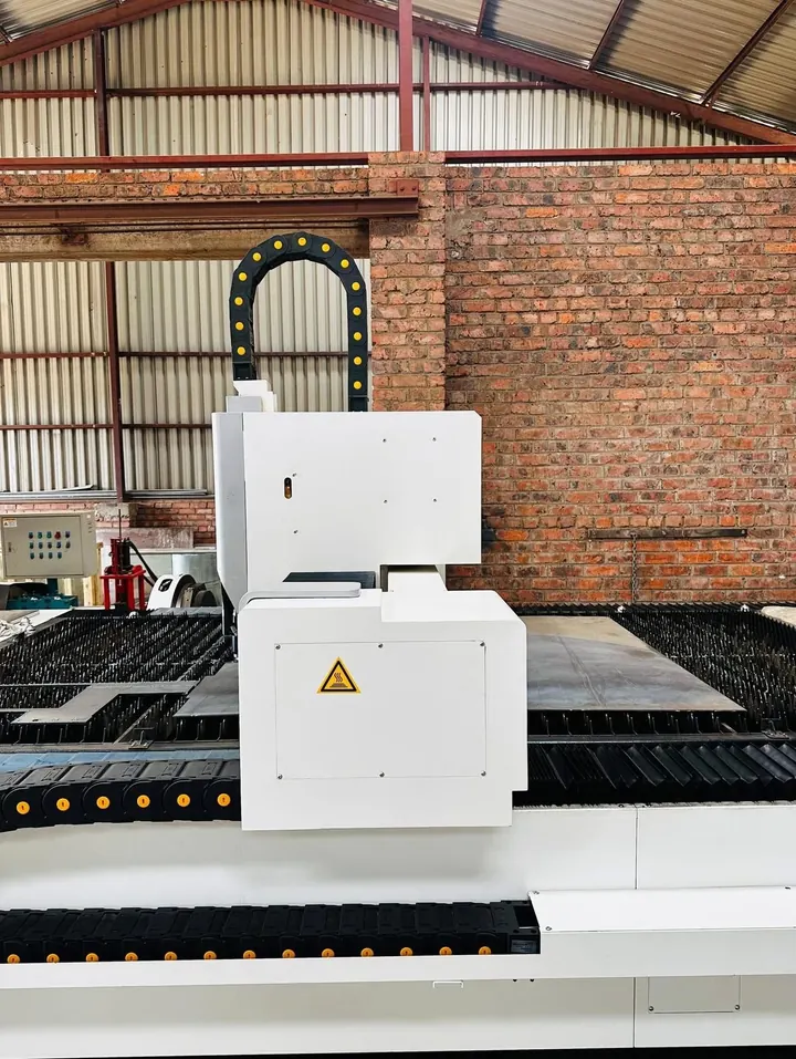 The laser gantry carriage seen side-on, cable chain arching over it, travelling above a slatted bed loaded with steel plate, with a brick wall and workshop sheeting behind.