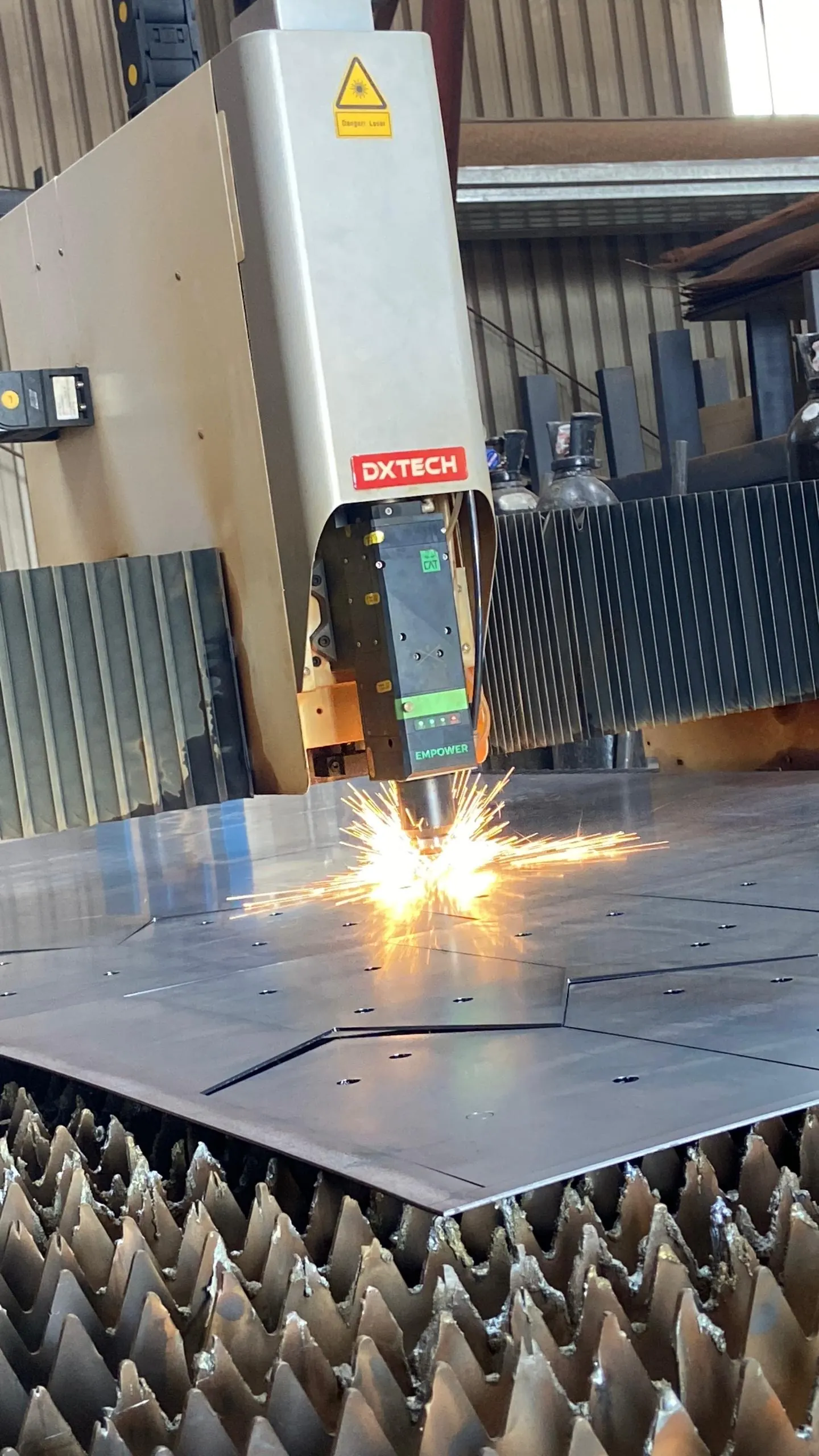 A DXTECH fibre laser cutting steel sheet, sparks thrown from the nozzle across the plate, with the slatted bed below and gas cylinders against the workshop wall behind.