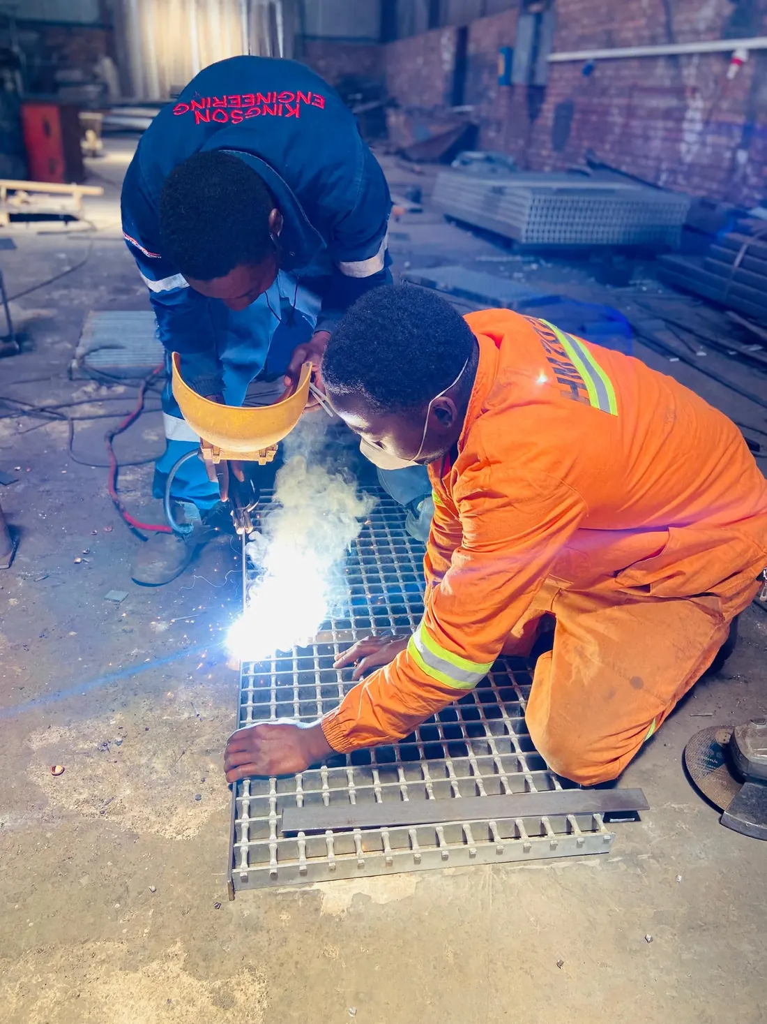 Two workers crouched over a steel grating panel, one holding a welding shield and striking an arc, the other steadying the panel; the overalls are lettered KINGSON ENGINEERING.